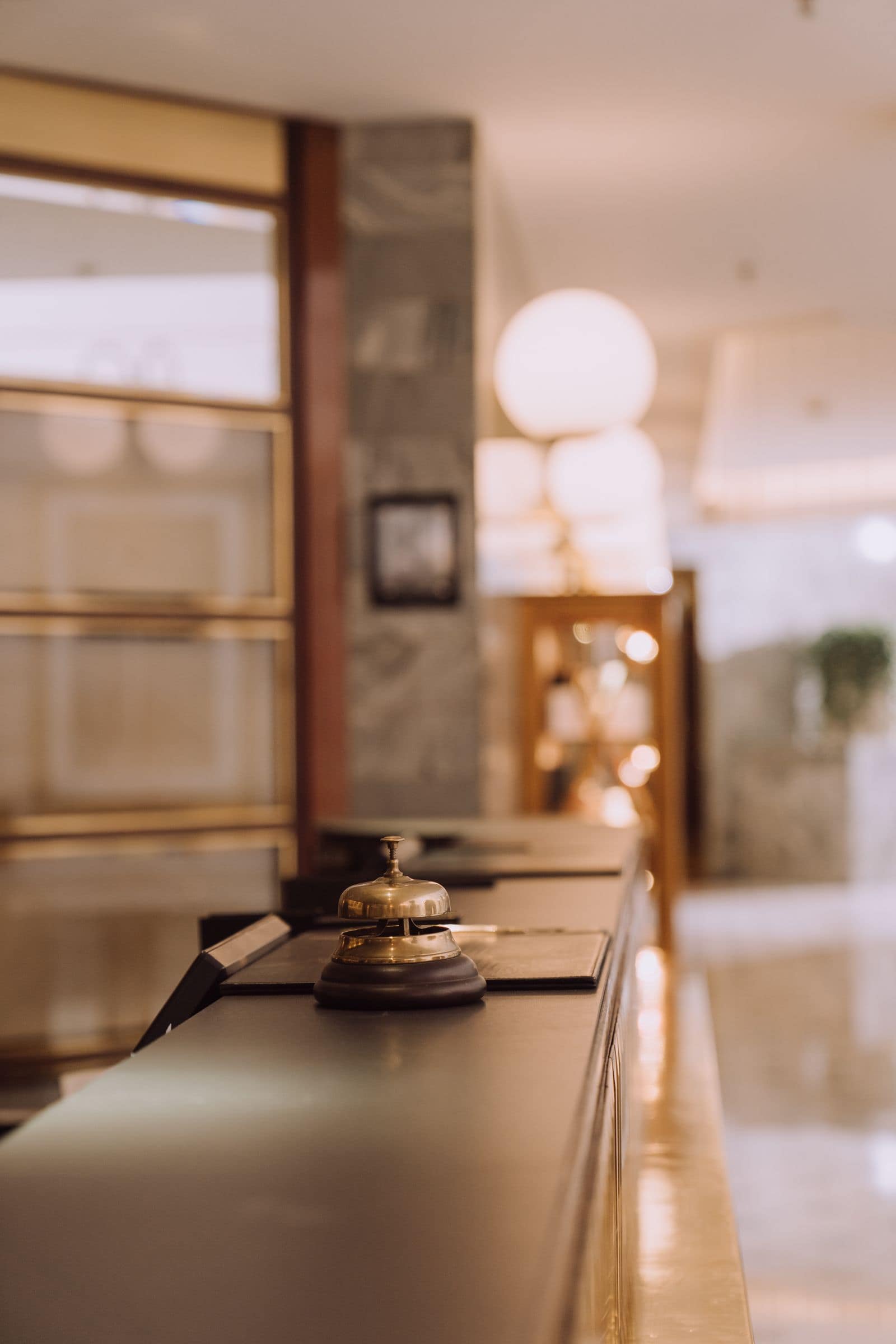 A brass service bell on a hotel reception desk, warm lamps glowing behind. Photo via Pexels (photo 5371683).